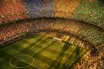 Overhead aerial view of a floodlit stadium with colourful group-section stands and crowd atmosphere under golden evening lights.