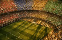 Overhead aerial view of a floodlit stadium with colourful group-section stands and crowd atmosphere under golden evening lights.