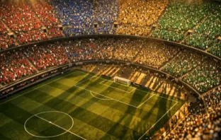 Overhead aerial view of a floodlit stadium with colourful group-section stands and crowd atmosphere under golden evening lights.