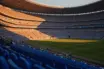 Floodlit football stadium at golden-hour kick-off with packed stands, dramatic shadows on empty pitch, Argentina and Egypt national colours.
