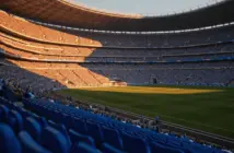 Floodlit football stadium at golden-hour kick-off with packed stands, dramatic shadows on empty pitch, Argentina and Egypt national colours.
