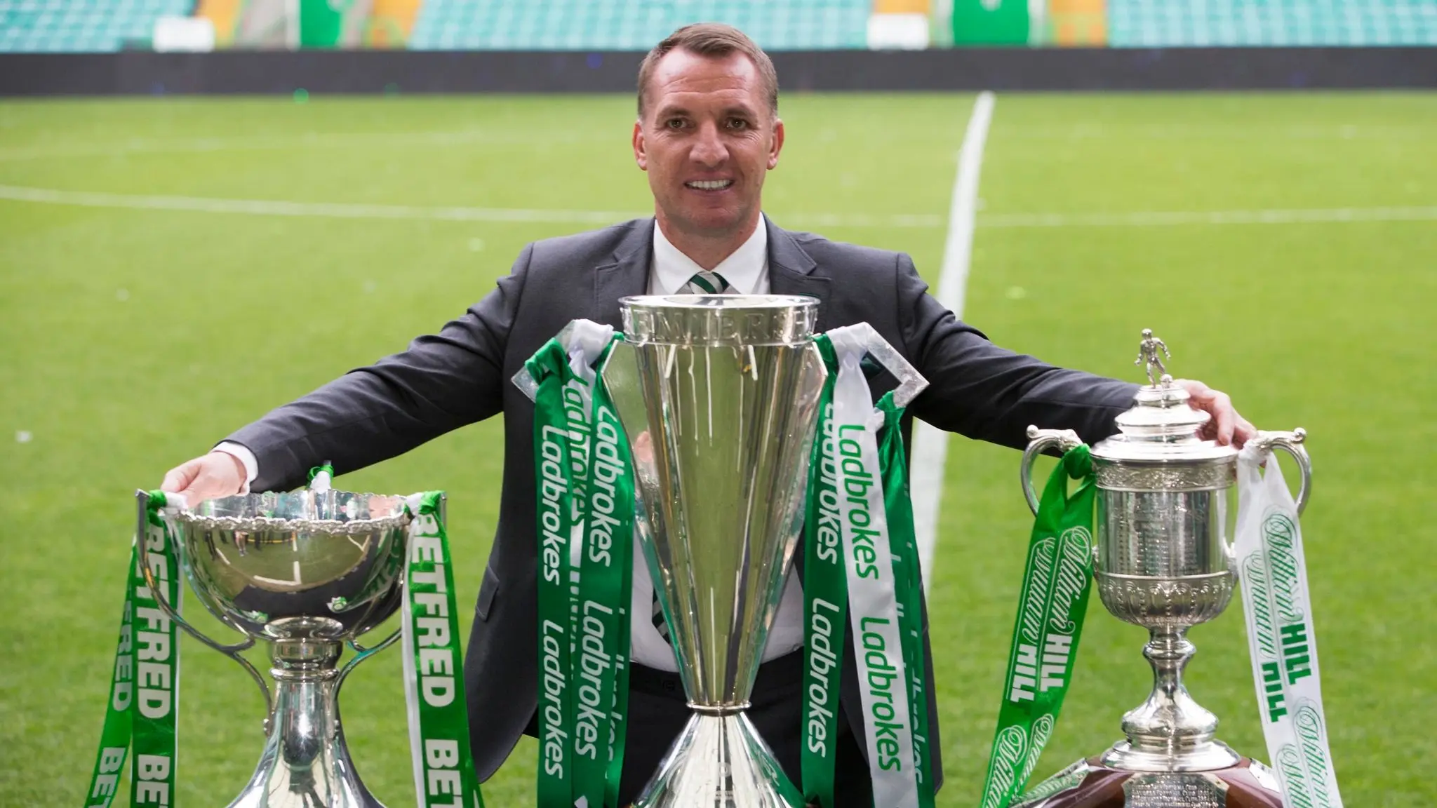 Brendan Rodgers holding trophies on a soccer field with Celtic FC branding.