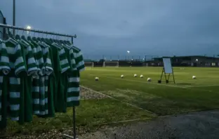 Empty Celtic training ground at dusk with unused hooped jerseys on rack, evoking summer transfer uncertainty