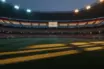 Floodlit stadium with packed stands and dramatic shadows on empty pitch at golden-hour dusk.
