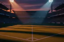 Empty floodlit football pitch at dusk with stadium stands in shadow, dramatic lighting creating geometric patterns.