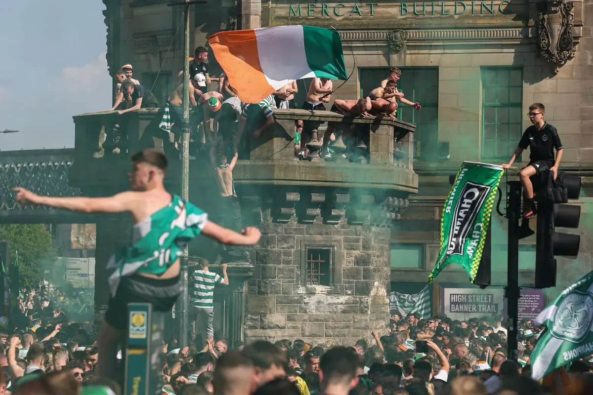 Celtic supporters celebrating on a balcony and in a crowd near the Mercat Building with an Irish flag.