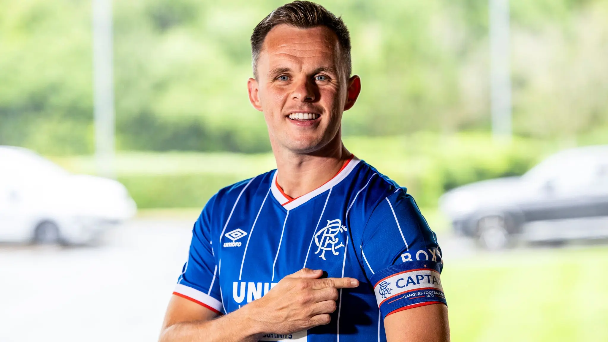 A man wearing a blue Rangers FC jersey with a captain's armband, pointing to the club crest.