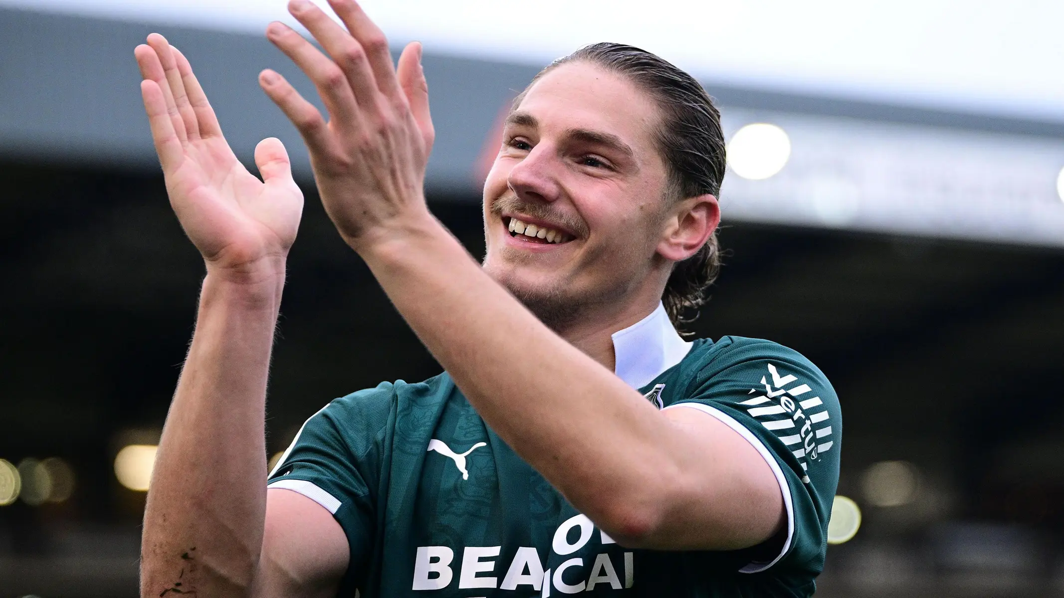 A football player in a green kit smiling and applauding fans.