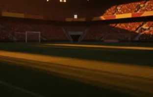 Floodlit stadium at golden hour with packed stands, dramatic shadows across empty pitch. Burgundy, green, red and gold colours in crowd.