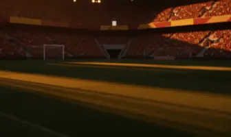 Floodlit stadium at golden hour with packed stands, dramatic shadows across empty pitch. Burgundy, green, red and gold colours in crowd.