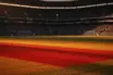 Floodlit stadium pitch at dusk with national flag colour tones and dramatic shadow patterns, no players visible.