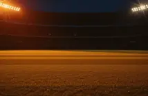Empty floodlit stadium pitch at golden hour with dramatic shadows, warm amber and red lighting, packed stands silhouetted against night sky.