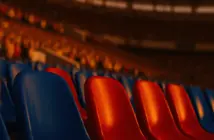 Close-up of blue and red stadium seats under golden-hour floodlights, packed stands in atmospheric bokeh.