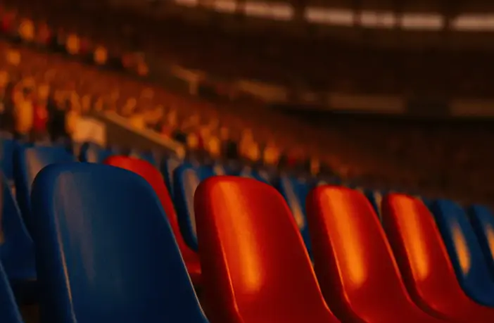 Close-up of blue and red stadium seats under golden-hour floodlights, packed stands in atmospheric bokeh.