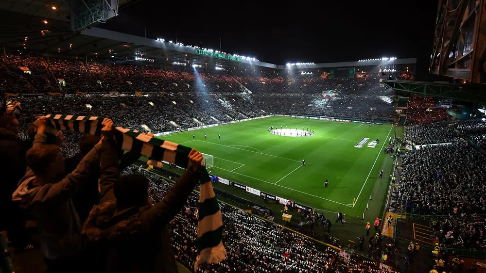 Wide shot of Celtic Park stadium in Glasgow at night with fans holding up a green and white striped scarf