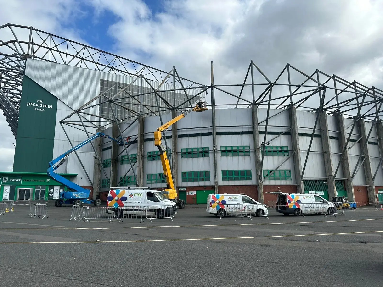Exterior view of the Jock Stein Stand at Celtic Park stadium with cherry pickers and service vans present.