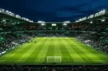 Floodlit football stadium at night with emerald green pitch and atmospheric green lighting in the stands