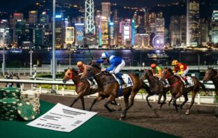 Thoroughbred racehorses galloping at a floodlit Hong Kong track with city skyline bokeh and betting chips on green felt