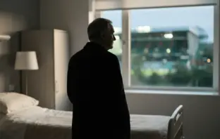 Respectful silhouette of a football manager near a hospital window with a softly lit stadium beyond