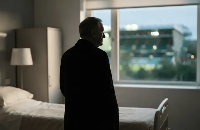 Respectful silhouette of a football manager near a hospital window with a softly lit stadium beyond