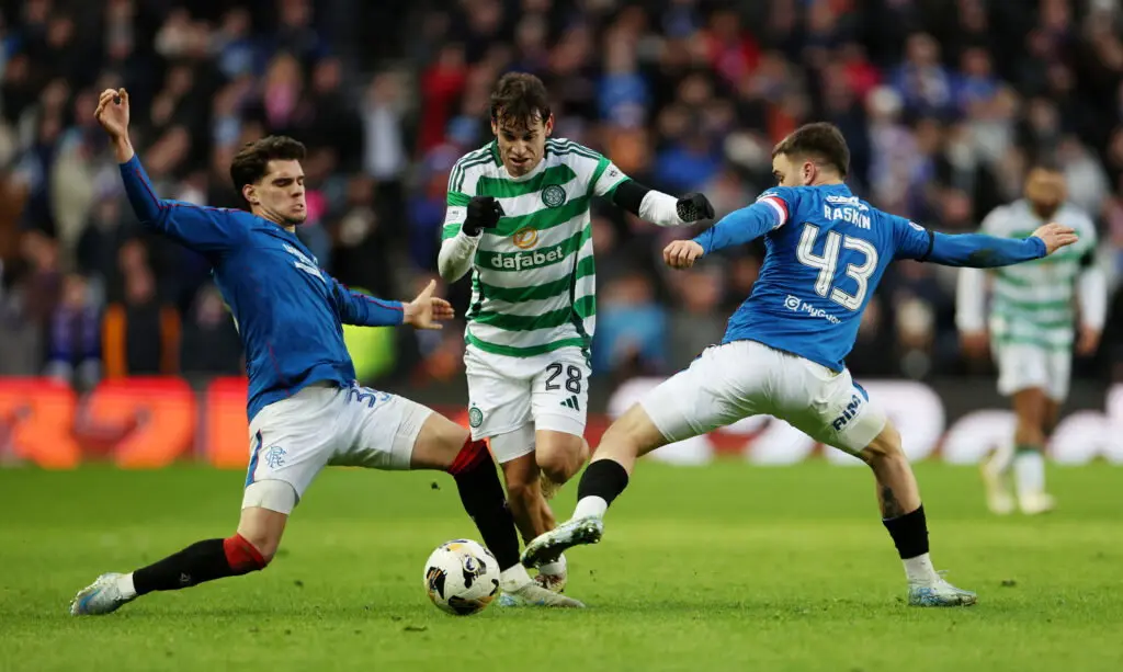 A Celtic player in a green and white hooped kit controlling the ball between two Rangers players in blue kits.