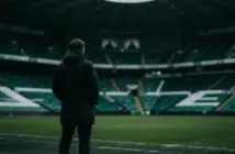 Non-identifiable football manager overlooking a green-lit stadium amid Celtic financial pressure