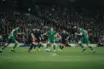 Cinematic football derby scene showing a green team battling for midfield control under stadium floodlights