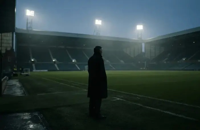 Silhouetted football manager facing an empty green pitch under stadium lights after a difficult run of defeats